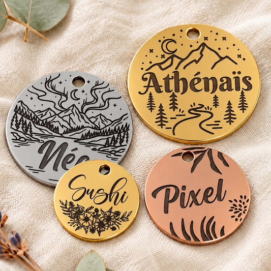 Quatre médailles personnalisées pour chiens en acier inoxydable doré, argenté et rose cuivré, gravées de prénoms et de motifs naturels, disposées sur un tissu beige clair avec des feuilles d’eucalyptus et des fleurs séchées. Medaléa